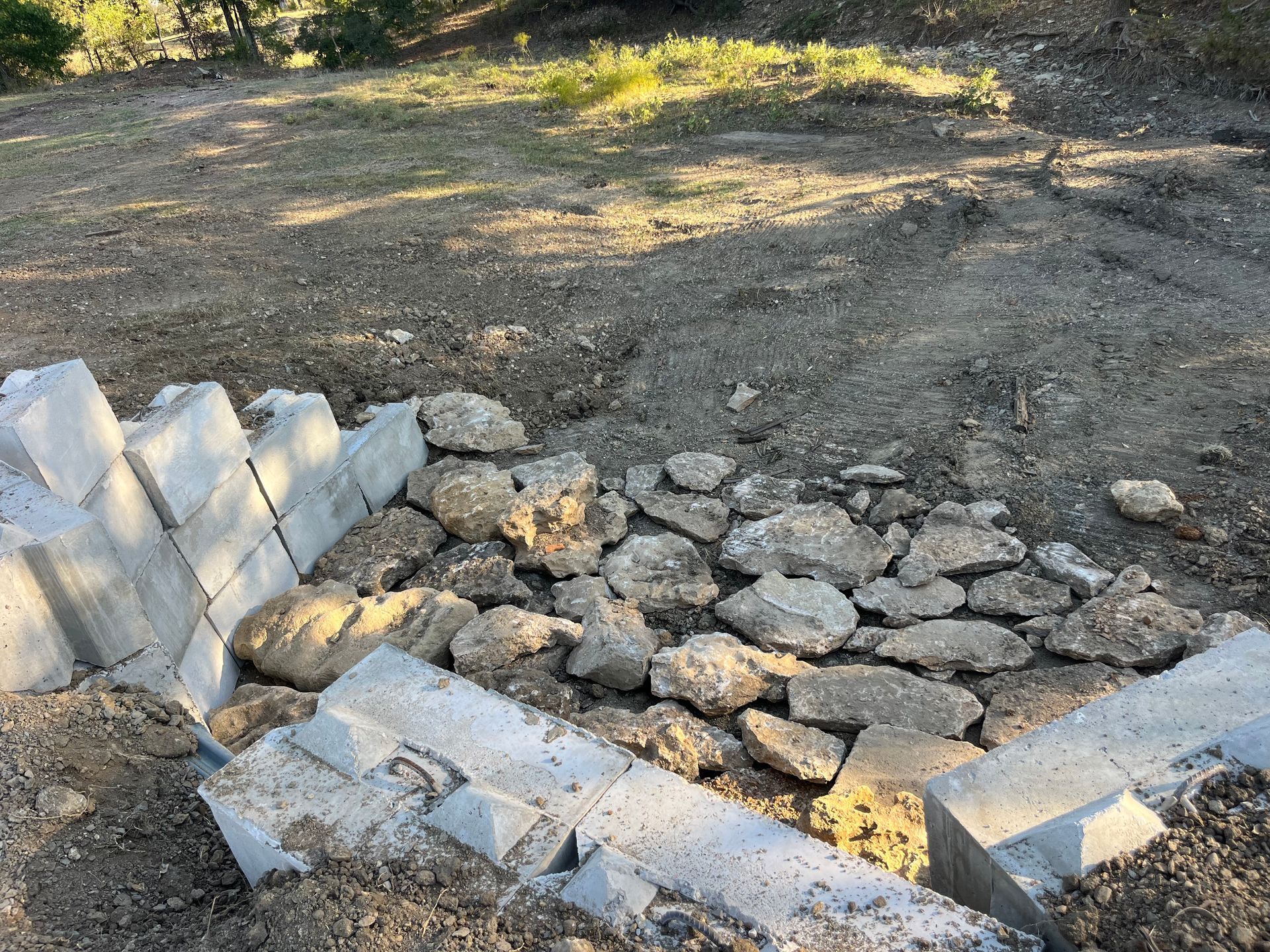 Construction site with concrete blocks and rocks; ground, hillside in background.
