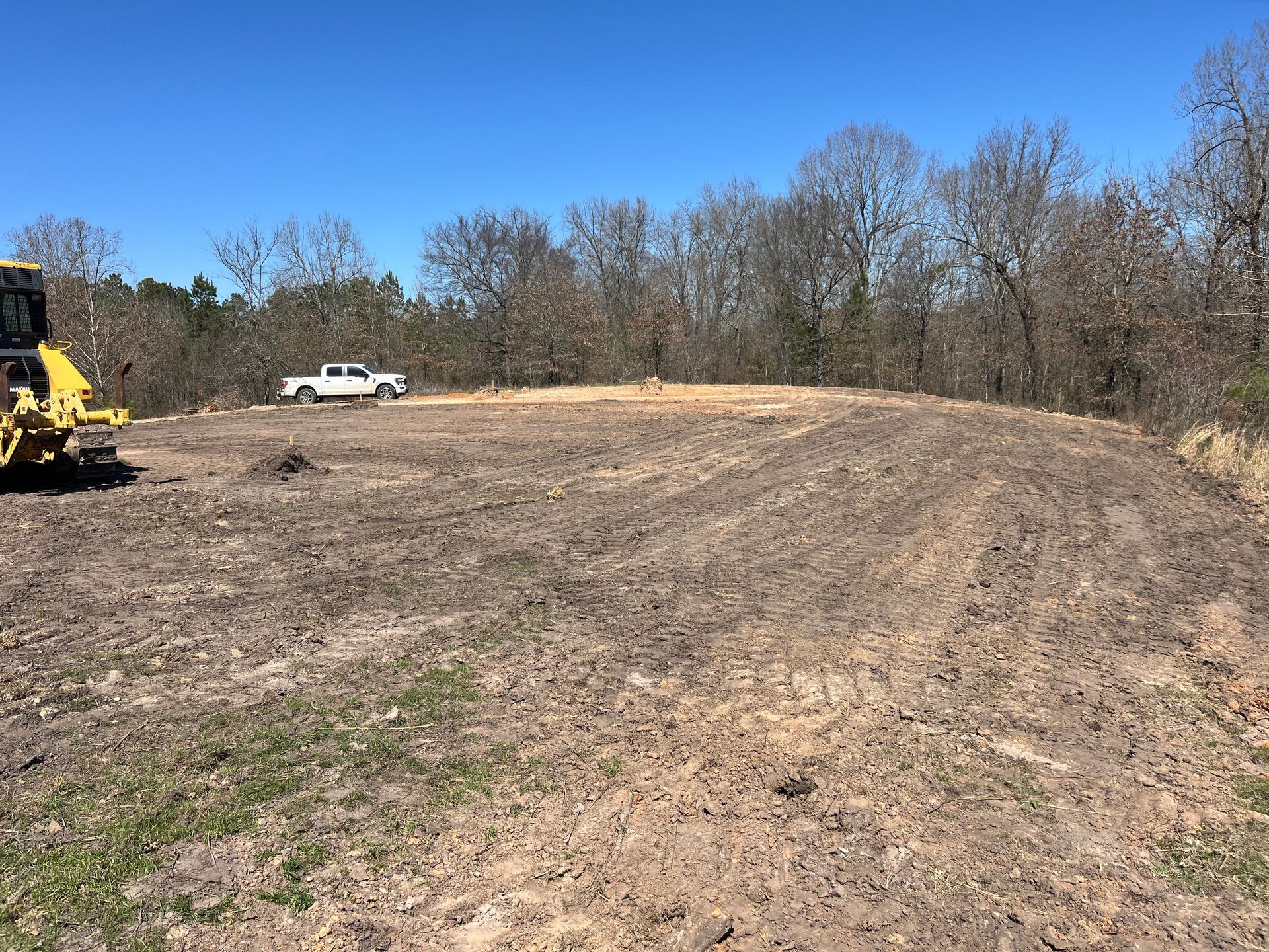A cleared land area with a truck and bulldozer, ready for construction. Brown dirt, blue sky, trees.