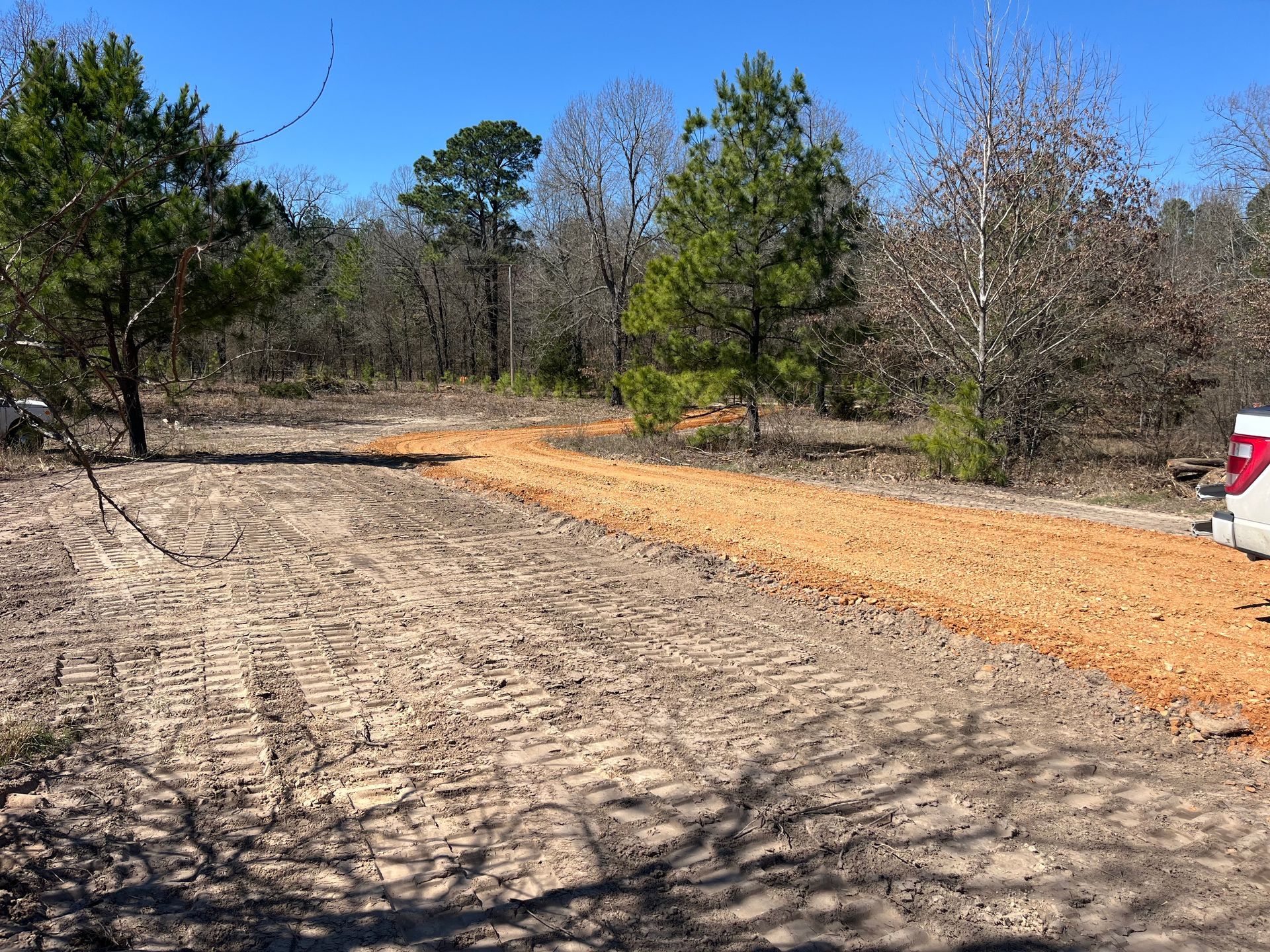 A dirt road winds through a wooded area, with a new layer of red gravel and tire tracks in the foreground.