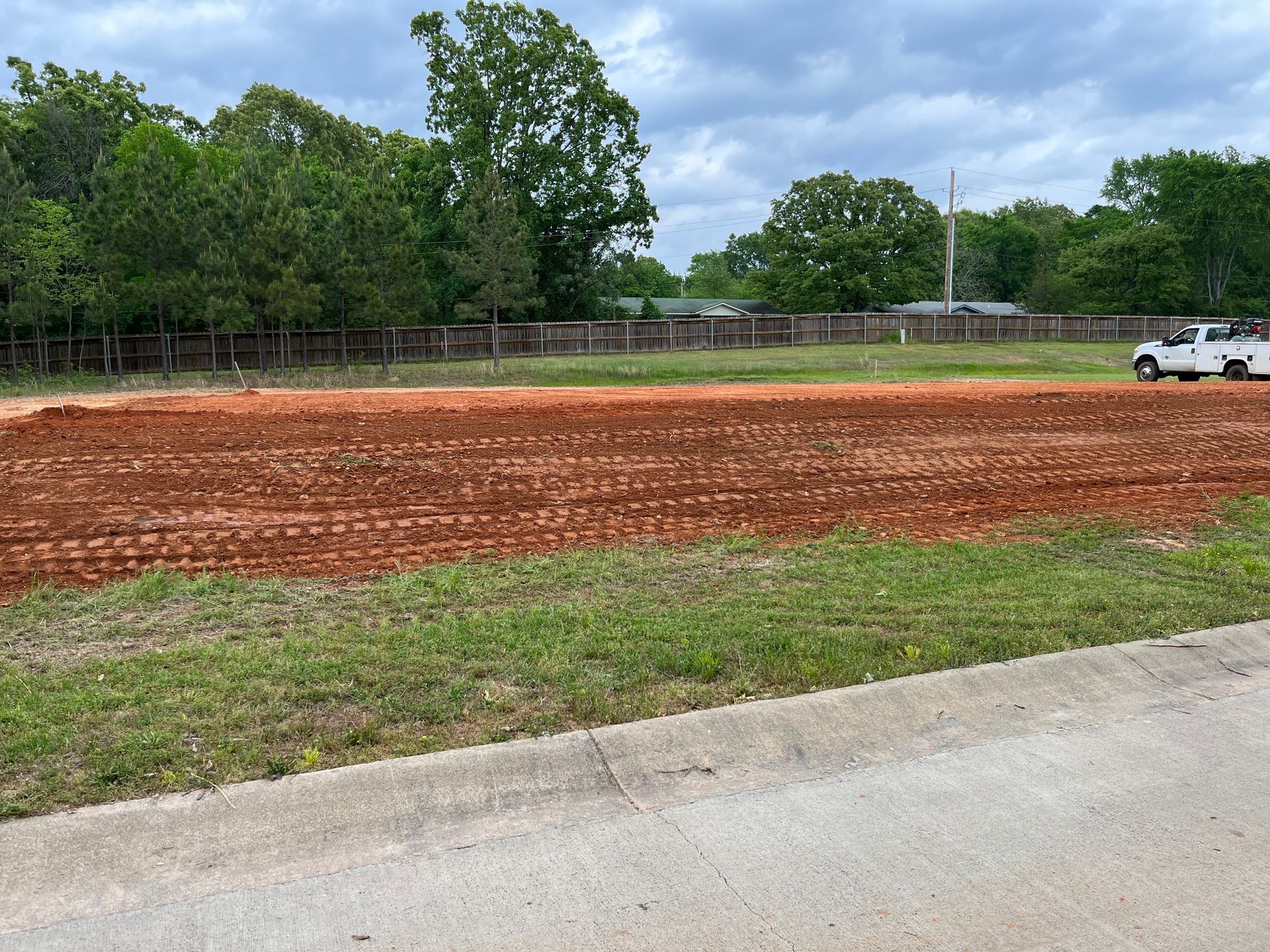 A red dirt lot with a truck, ready for construction, bordered by grass and a fence.