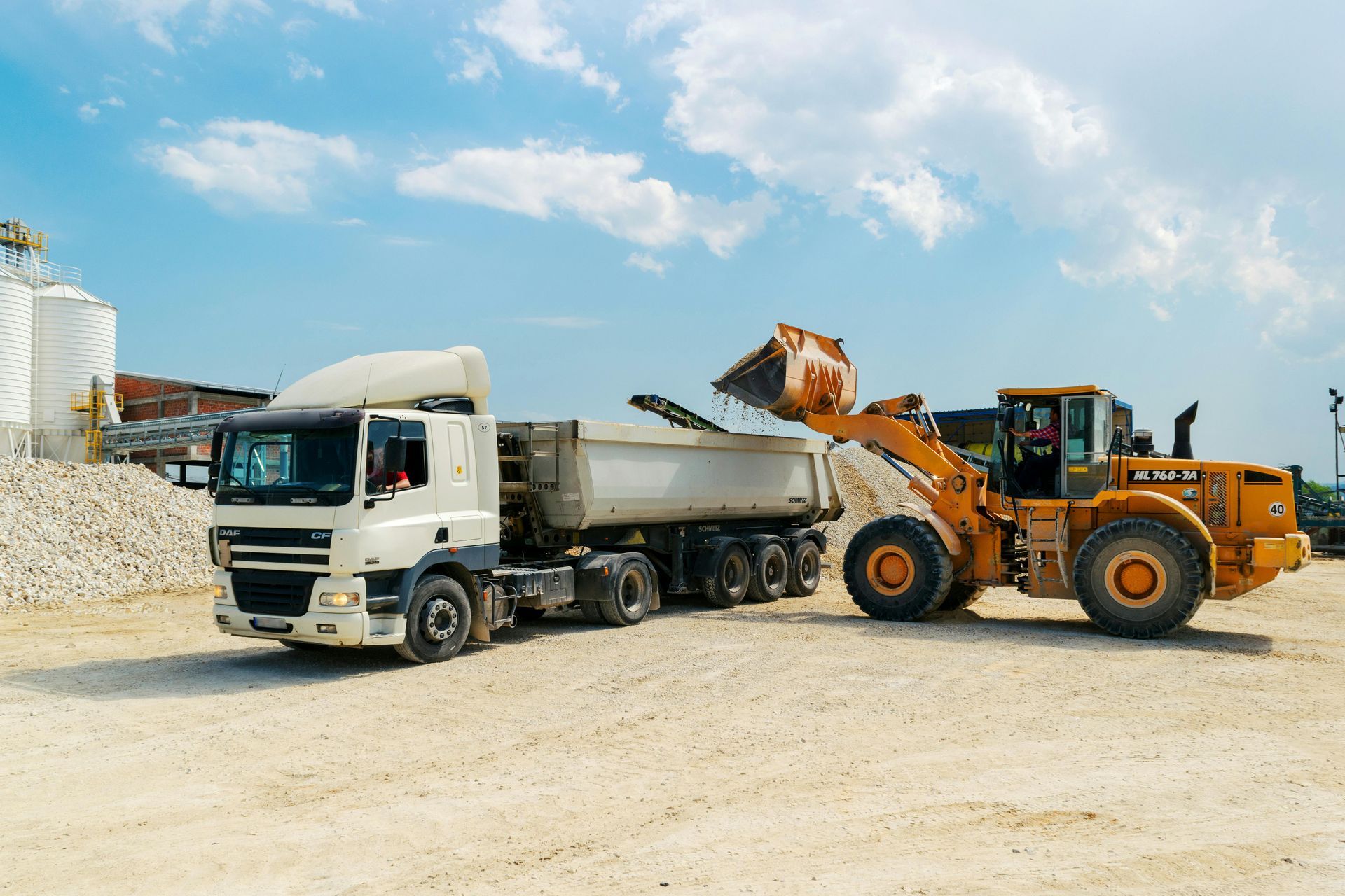Yellow front loader filling a truck with gravel under a blue sky at a construction site.