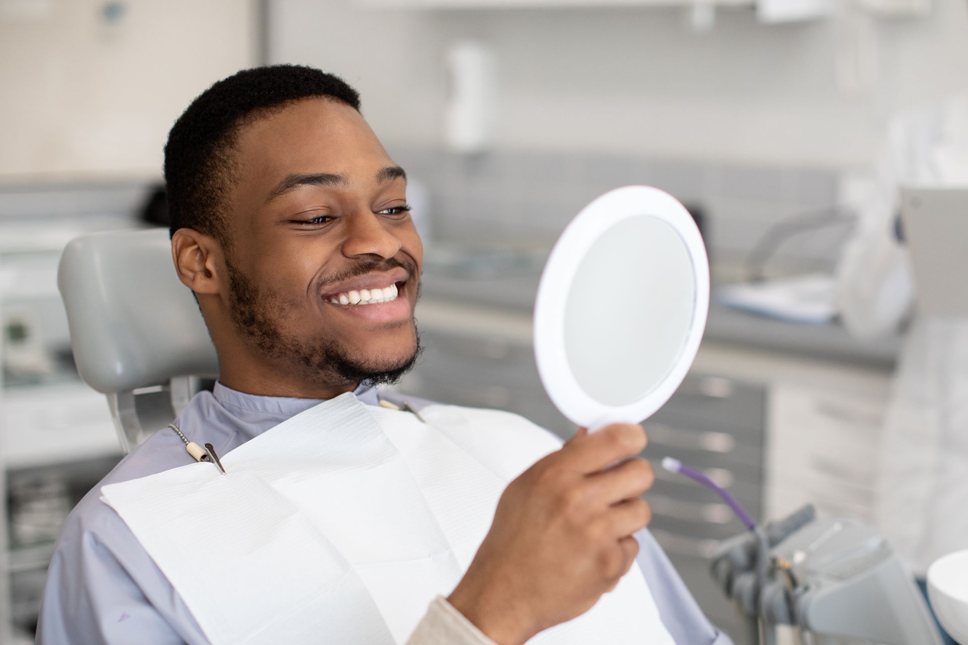 A man is sitting in a dental chair looking at his teeth in a mirror.