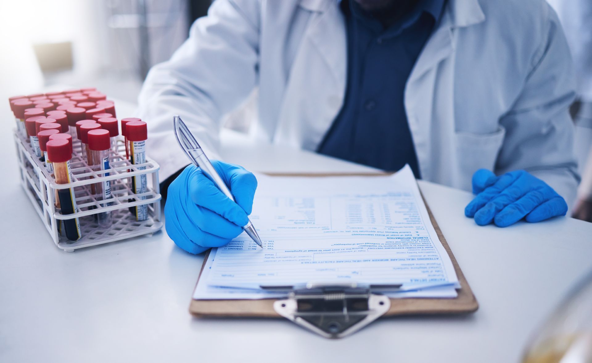 Scientist in lab coat and gloves writing on a clipboard next to a rack of blood sample tubes.