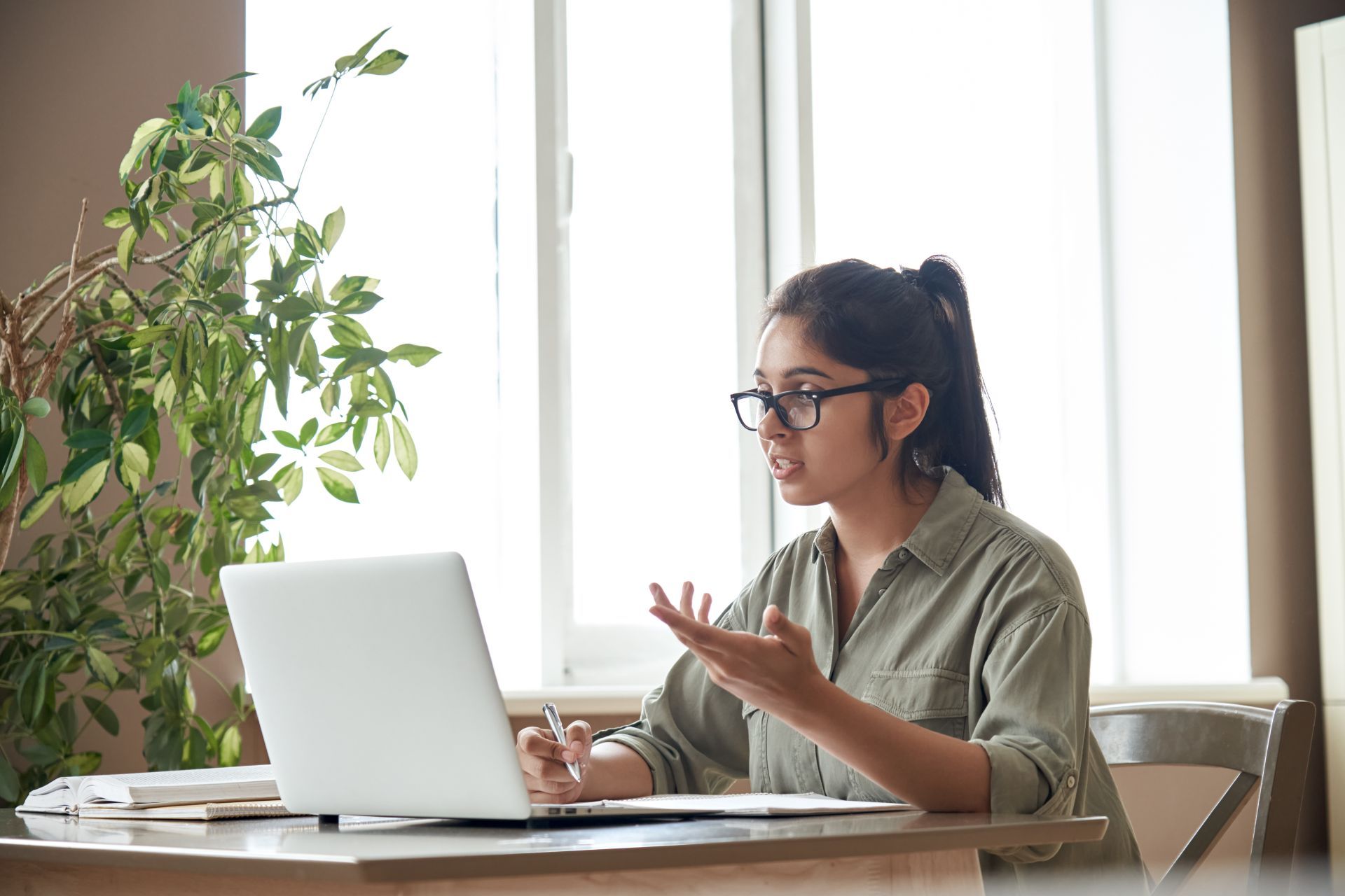 Woman in glasses, working on laptop, gesturing with hands, seated at a desk near a window.