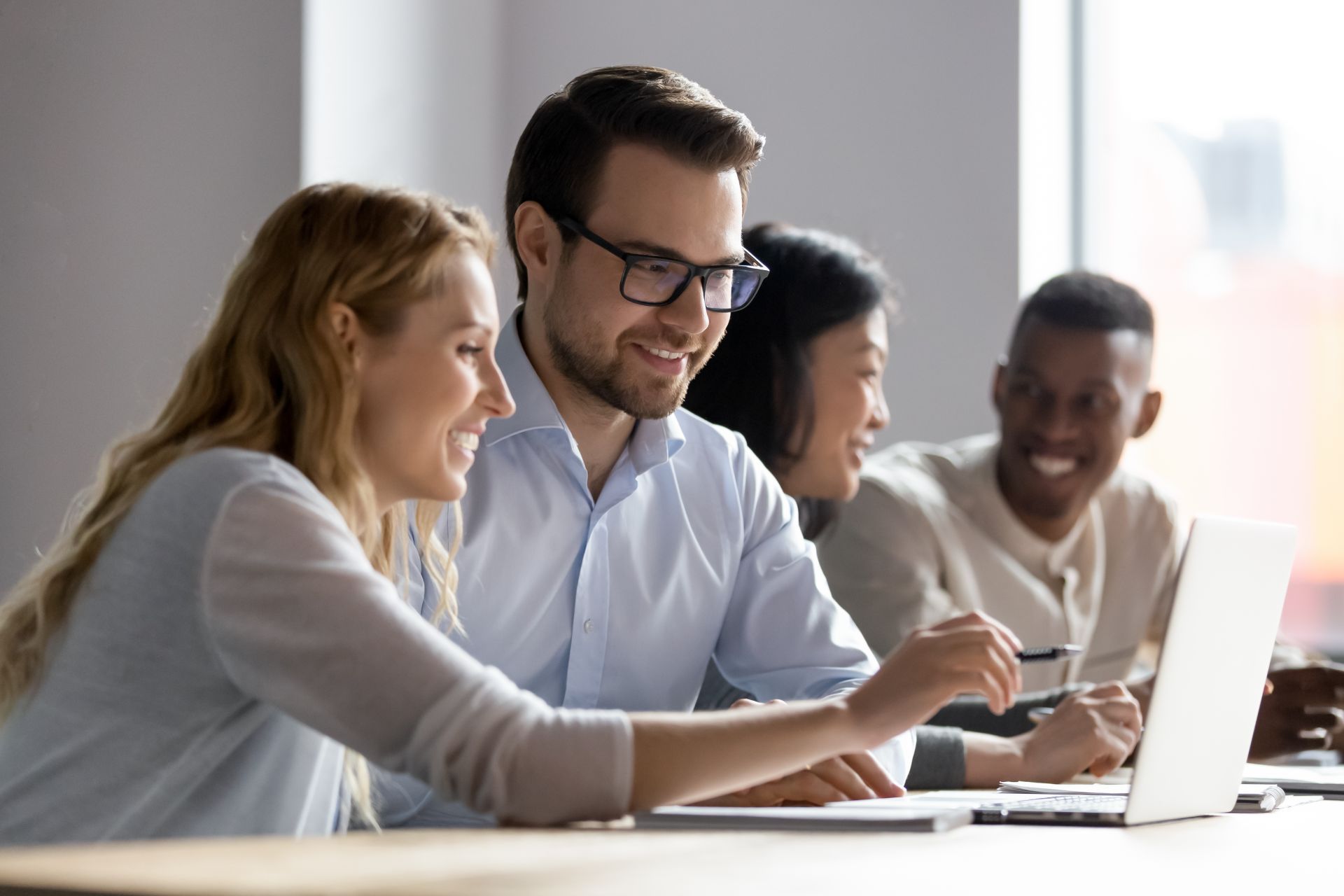 Four colleagues looking at a laptop, smiling.  One points at the screen. Office setting.