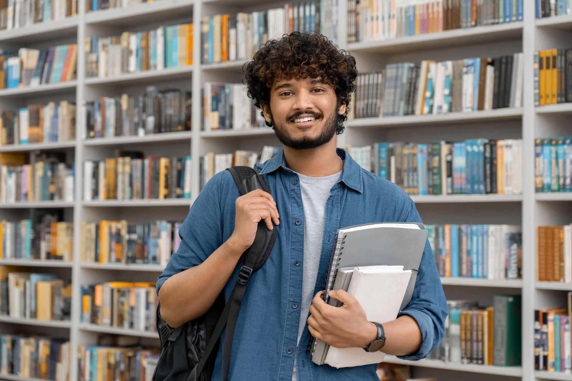 Man with curly hair smiling, holding books and a laptop in a library with bookshelves.
