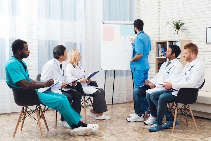 Doctors in a room, one presenting at a whiteboard, others seated, some taking notes, a plant in the background.