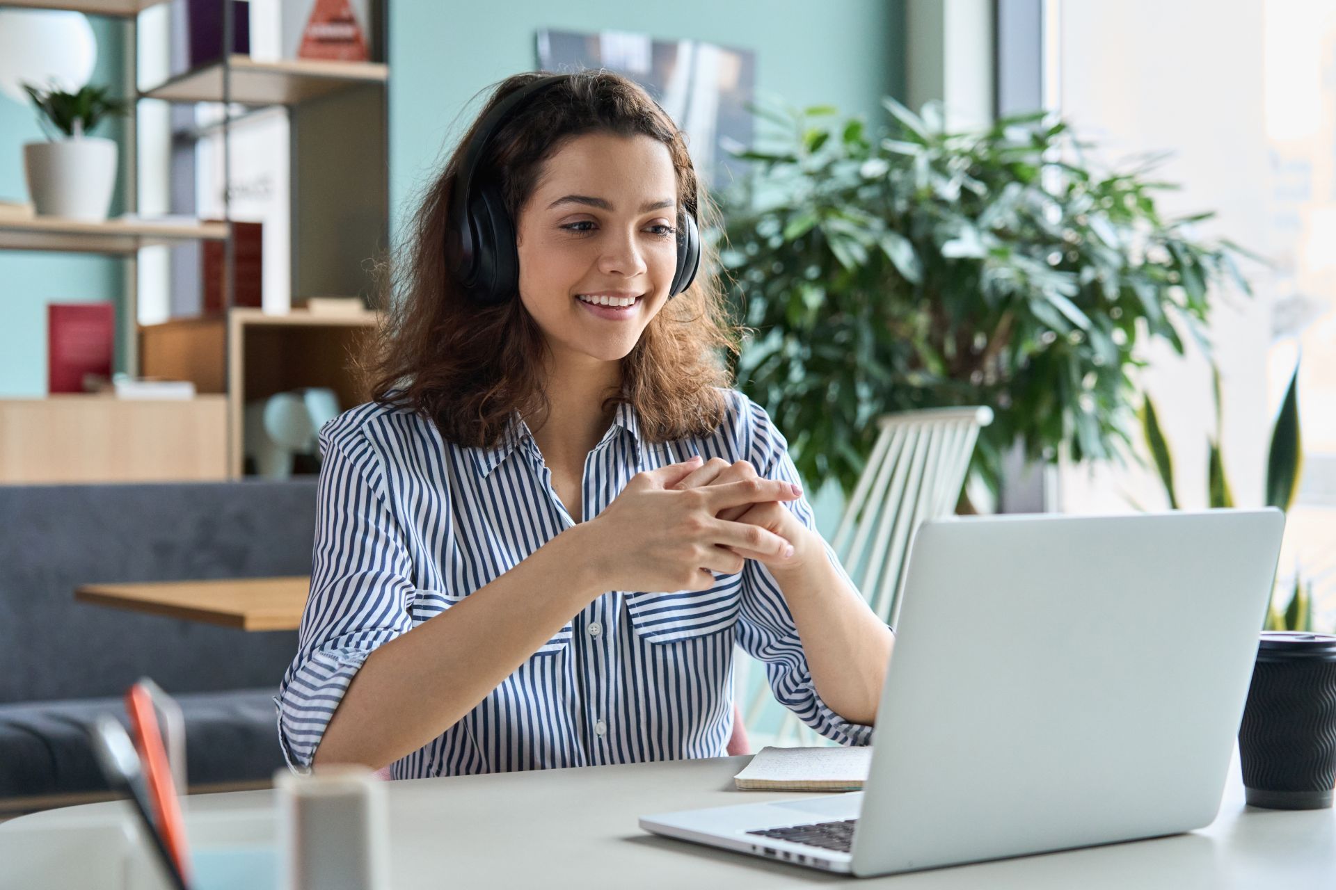 Woman wearing headphones, smiling, and gesturing while on a video call at a desk with a laptop.