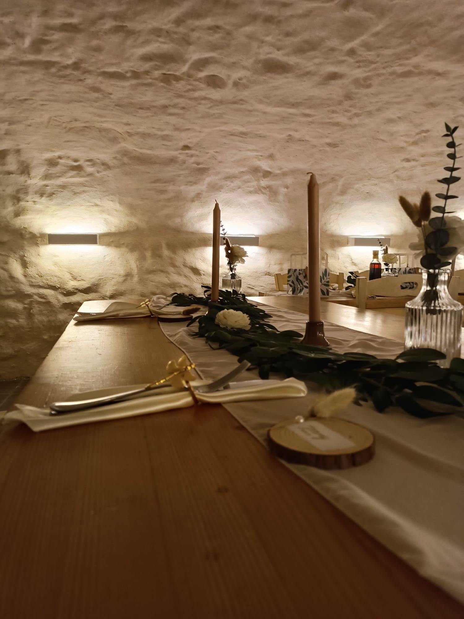 Ein rustikal gedeckter Esstisch aus Holz für eine Veranstaltung, mit Kerzen, Grünpflanzen und Tischkarten vor einer strukturierten Steinwand.