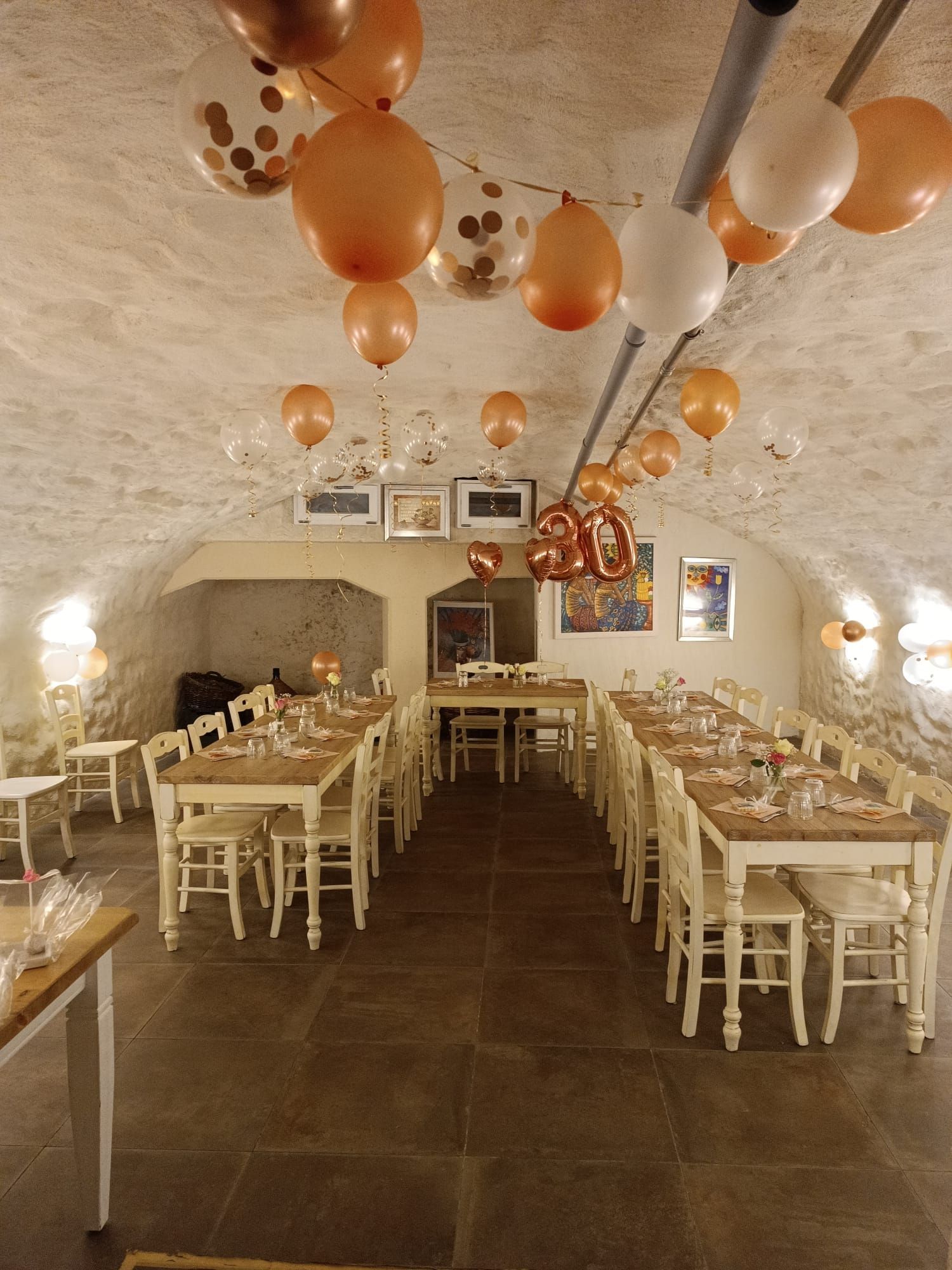 Ein steingewölbter Veranstaltungsraum, der für eine Party mit langen Holztischen, weißen Stühlen und hängenden goldenen und weißen Luftballons hergerichtet ist.