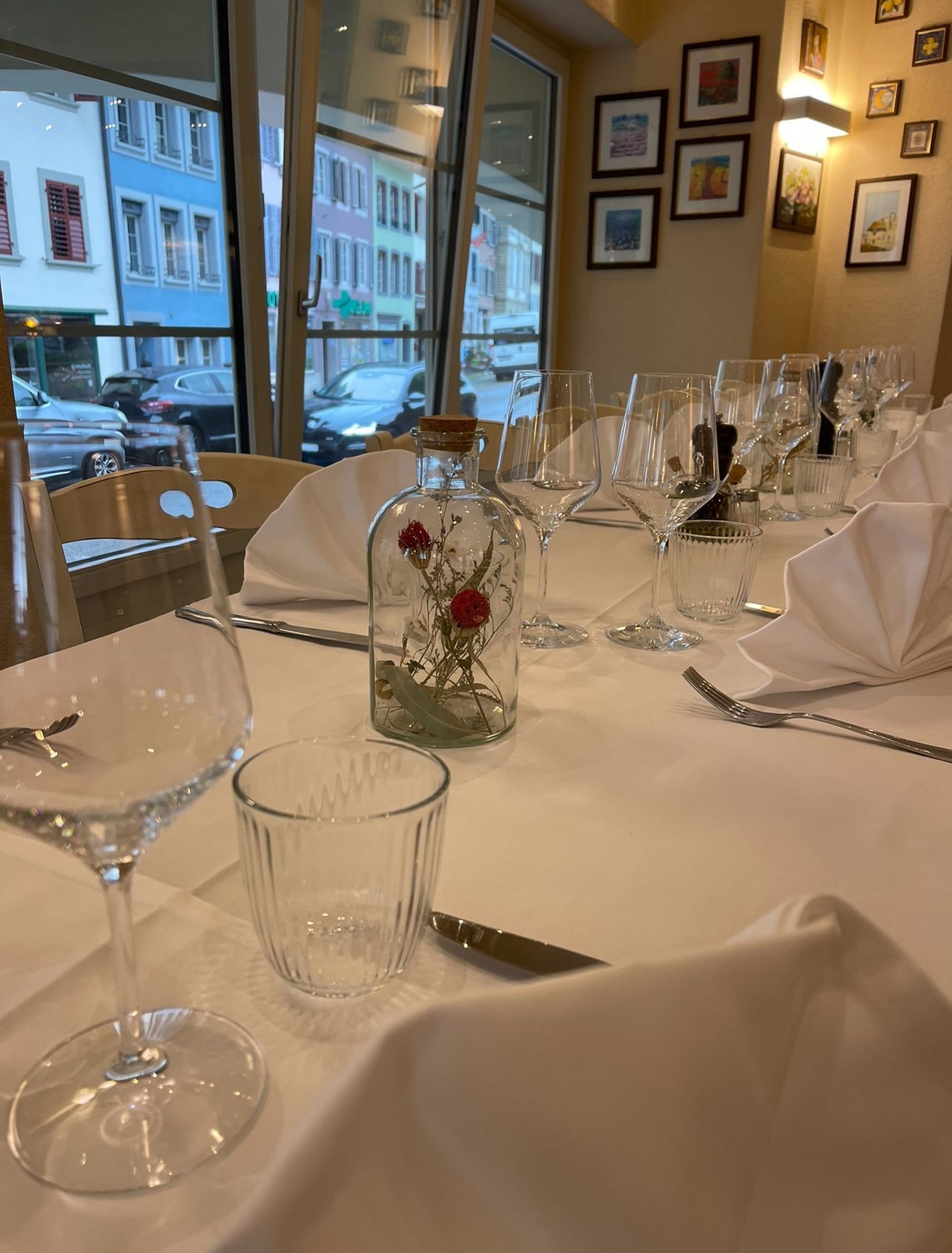 Ein zum Essen gedeckter Tisch mit weißen Tischdecken, Weingläsern, Servietten und einer kleinen Glasvase mit Blumen in der Nähe eines Fensters.