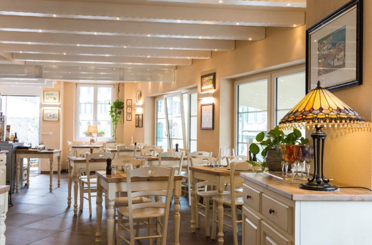 Innenansicht eines Esszimmers mit Holztischen, Stühlen, cremefarbenen Wänden, sichtbaren Deckenbalken und einer Tischlampe.