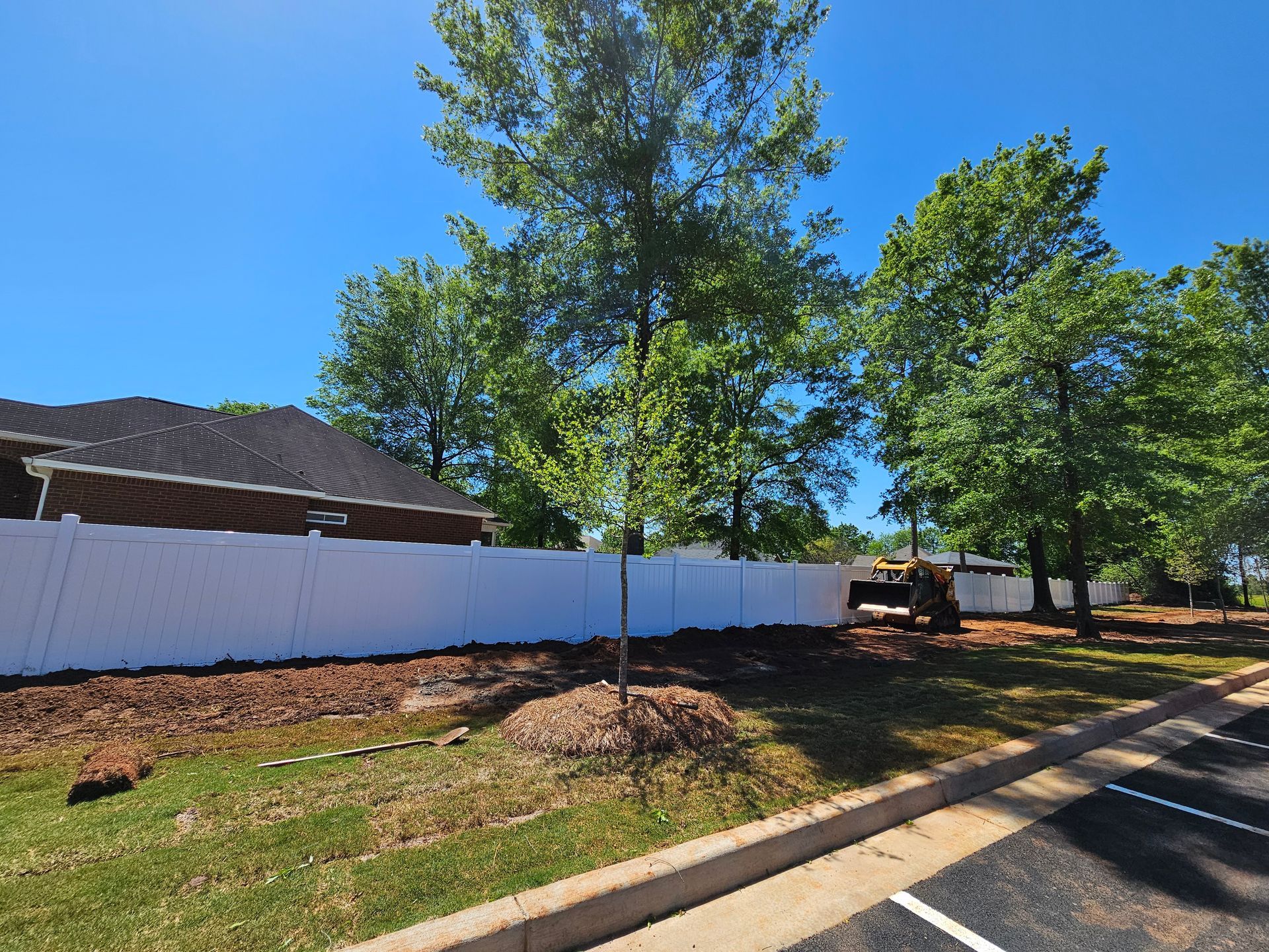 A white fence surrounds a yard with trees and a house in the background