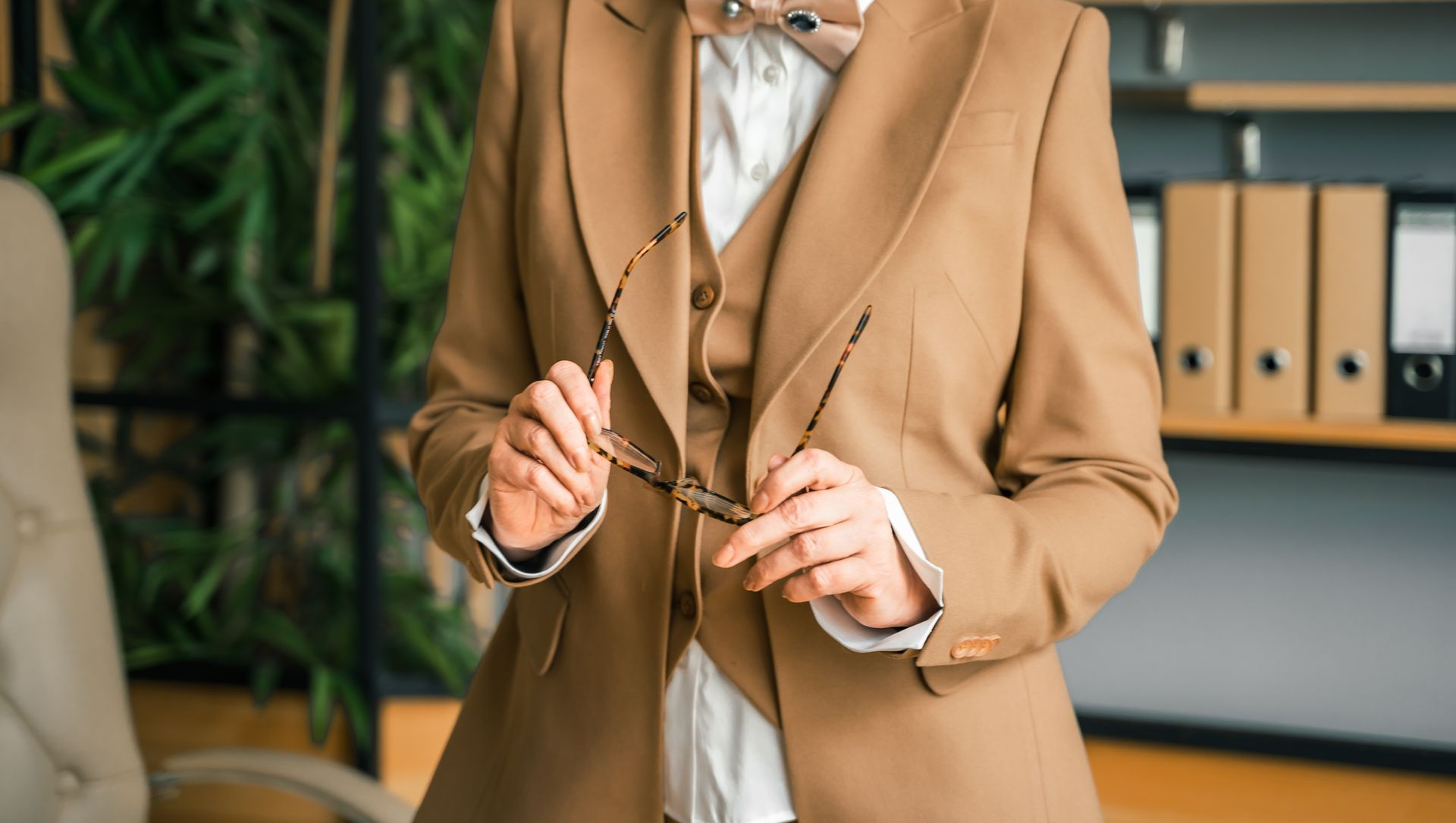 Woman in tan suit holding glasses. Office setting, folders on shelf.