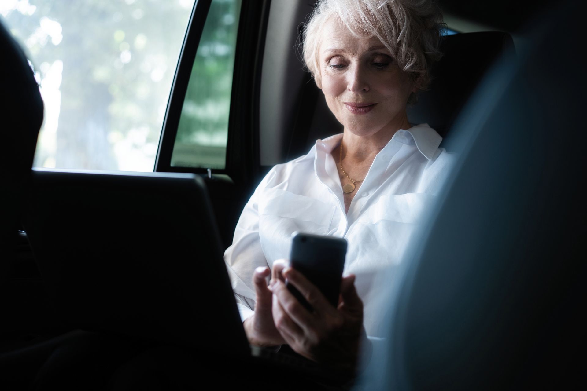 Woman in white shirt using a smartphone in a car. She is looking down and smiling.