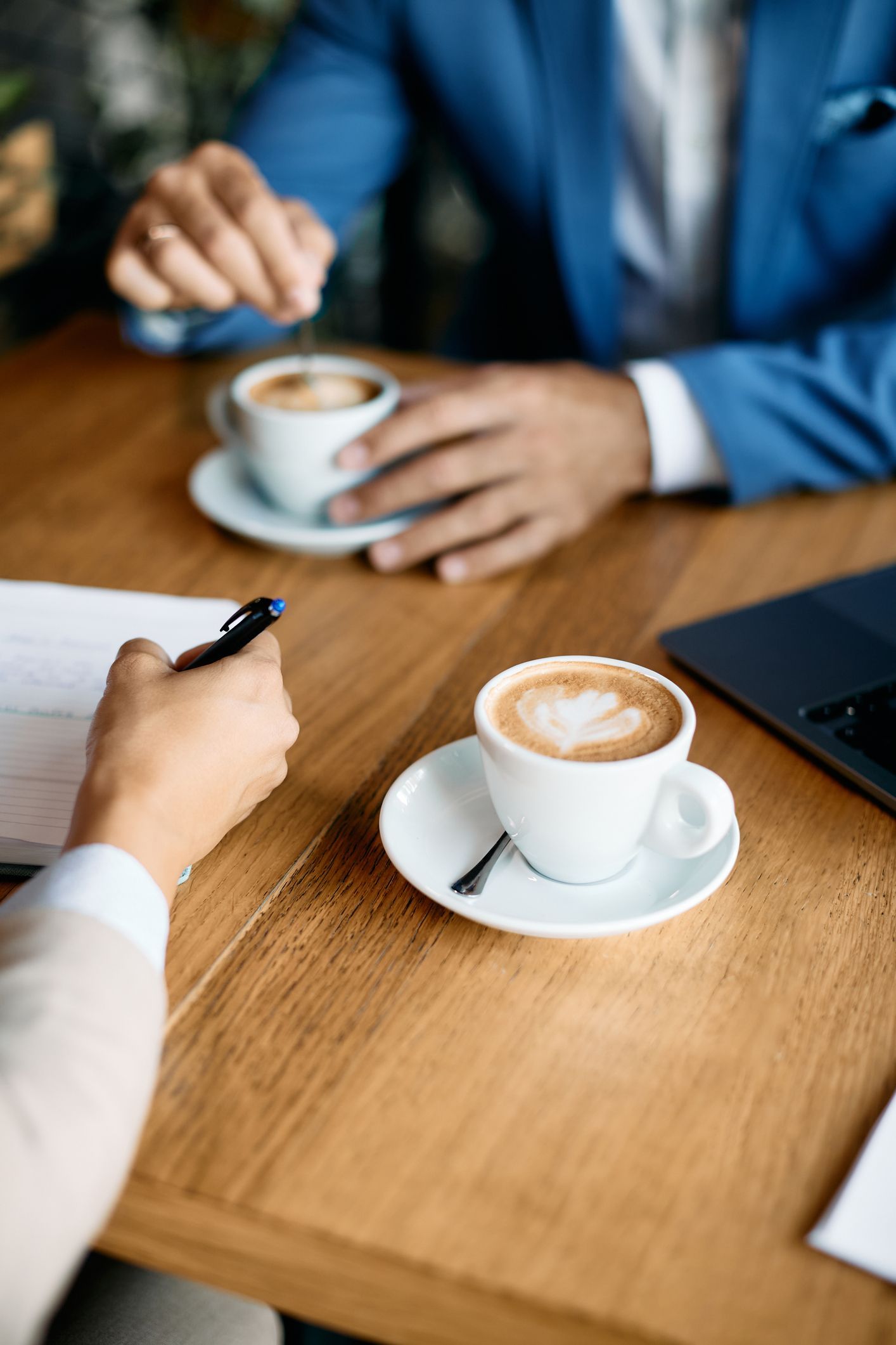 Two people at a wooden table, having coffee. One writes while the other gestures.