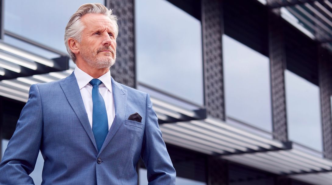 Man in blue suit, looking thoughtfully to the side, standing outside a modern building with glass and metal features.