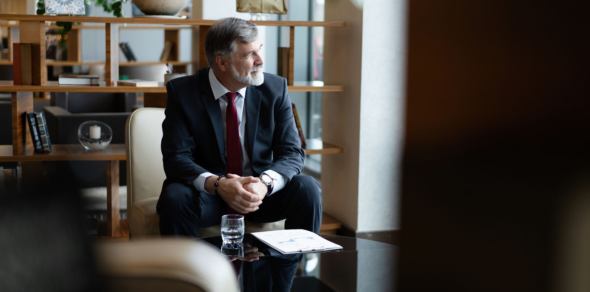 A man in a suit looks out a window. He sits with hands clasped in a modern office space.