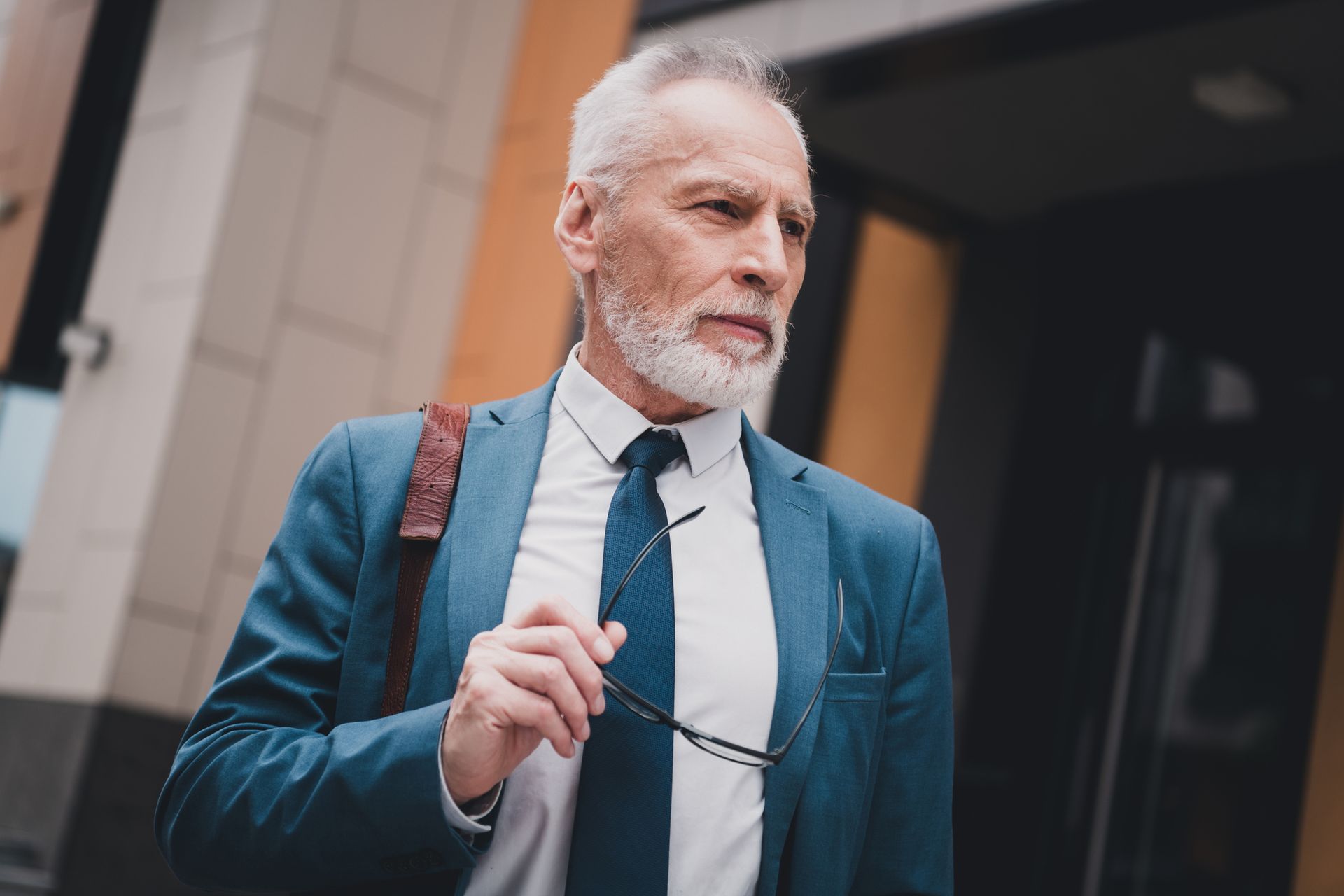 Older man in a blue suit holding glasses, looking thoughtful, outside a building.
