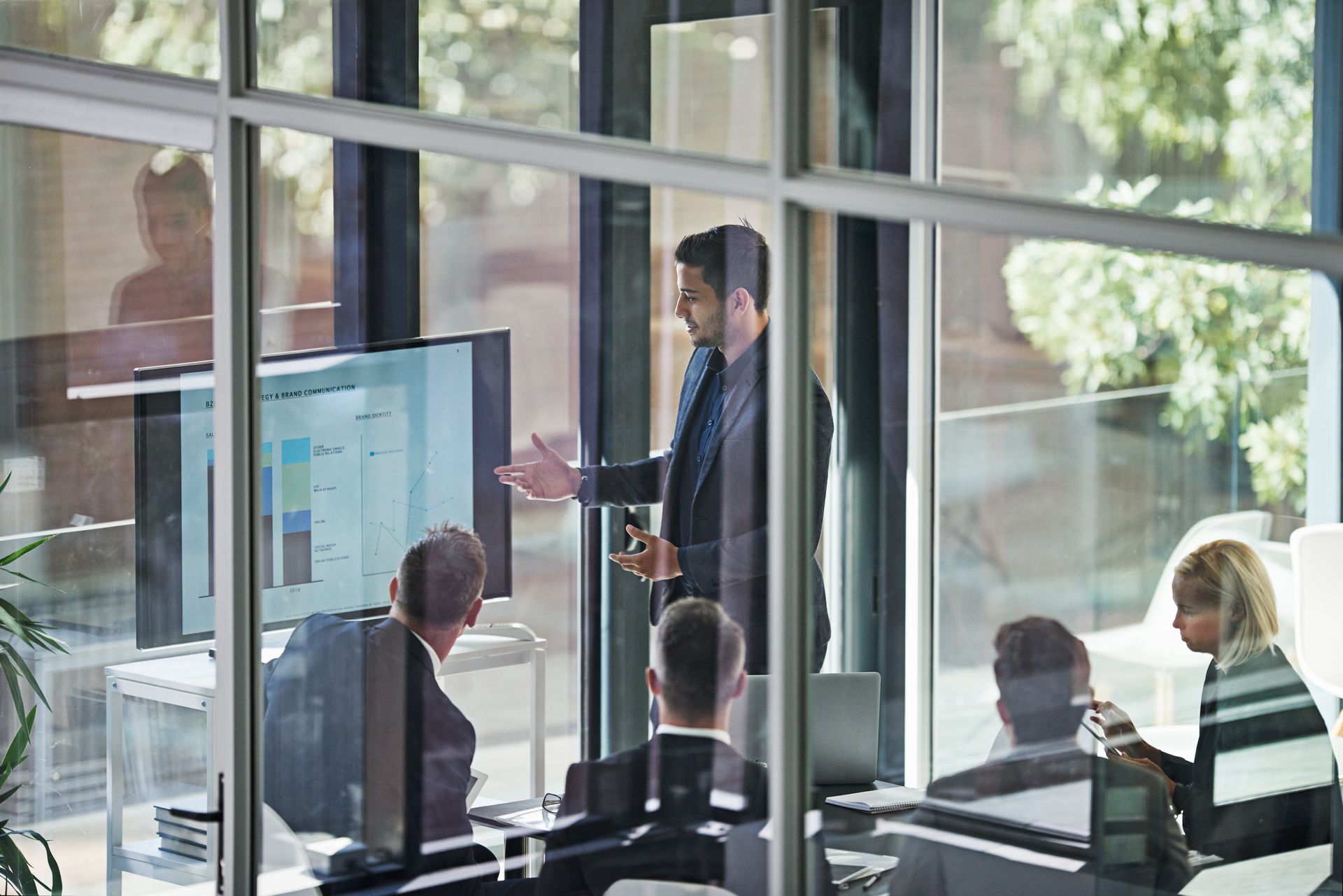 Man presenting data on screen to a team in a glass-walled office.