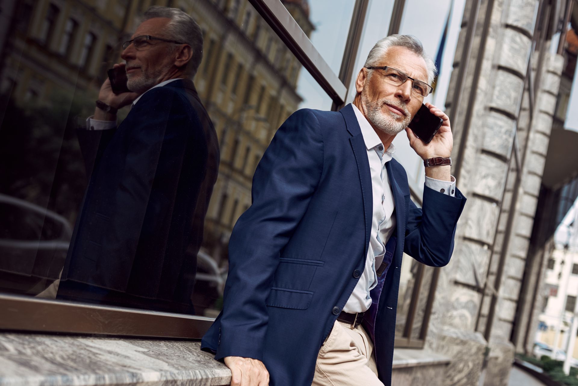 Man in blue blazer talks on the phone near a building, looking slightly to the side.