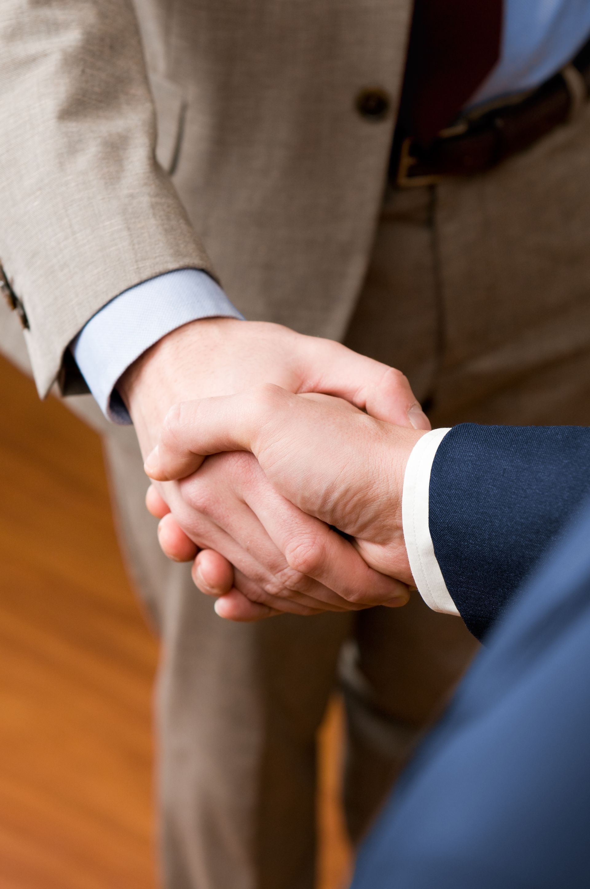 Two men in suits shaking hands, close up. Wooden floor visible in background.