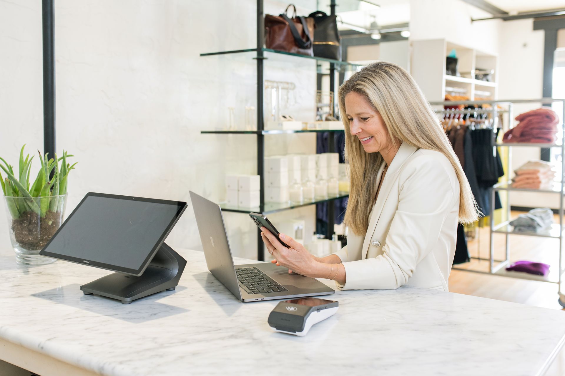 Woman smiles while looking at phone, laptop, and point-of-sale system on a store counter.