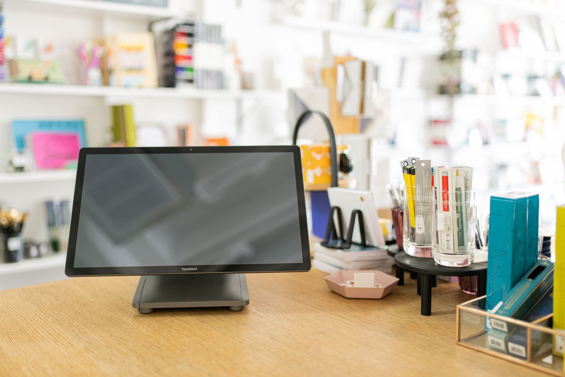 A point-of-sale touch screen display on a wooden counter in a store, with stationery items in the background.