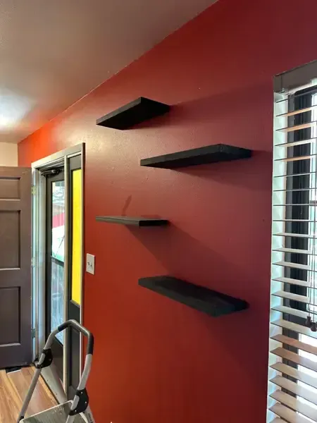 Red wall with four black floating shelves beside a window with blinds and an open door.
