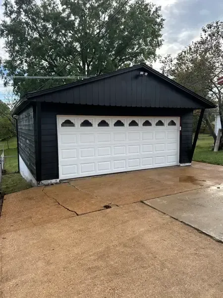 Black detached garage with white doors beside a wet concrete driveway and trees.
