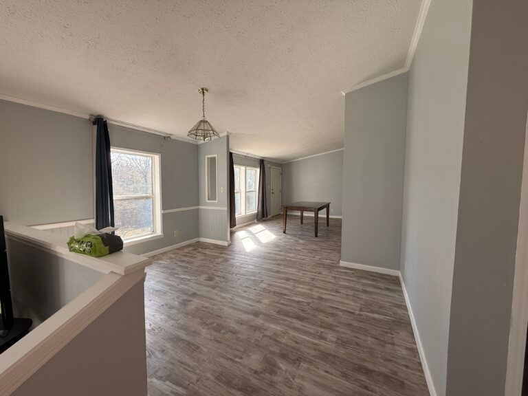 Empty gray living room with wood-look flooring, large windows, and a small dining table.