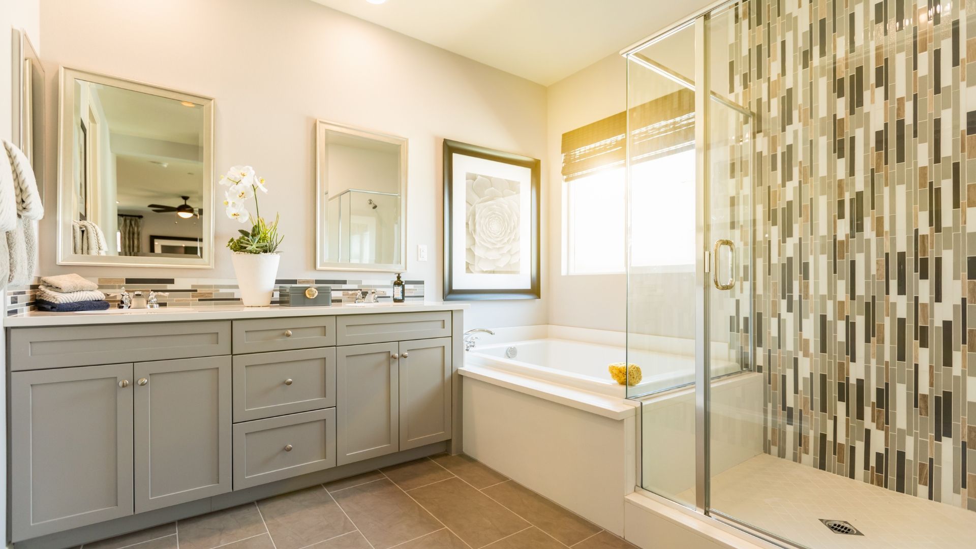 Bright bathroom with double vanity, mirrors, tub, and mosaic tile shower in beige and gray tones
