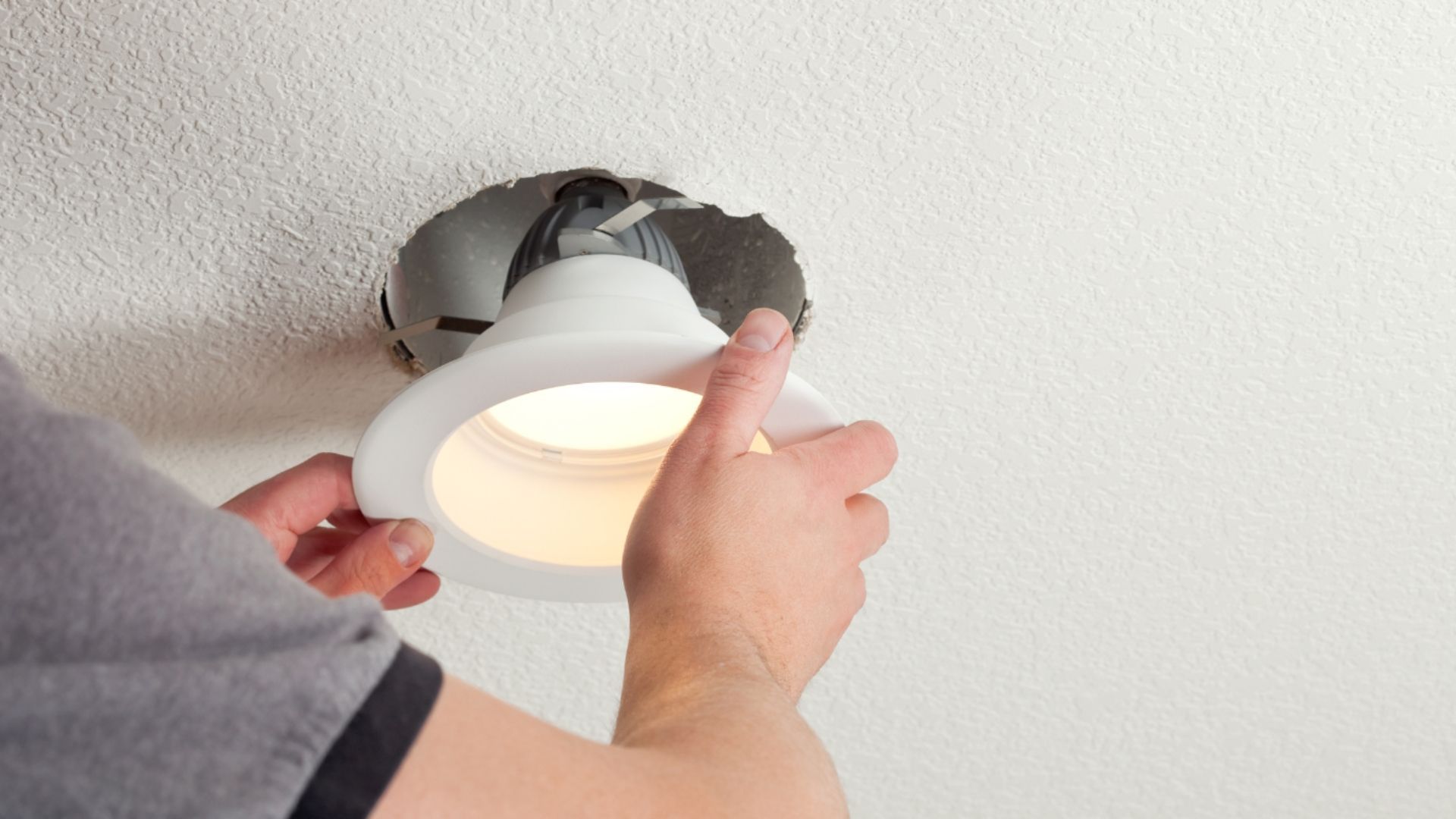 Hands installing a recessed ceiling light into a hole in the white ceiling.