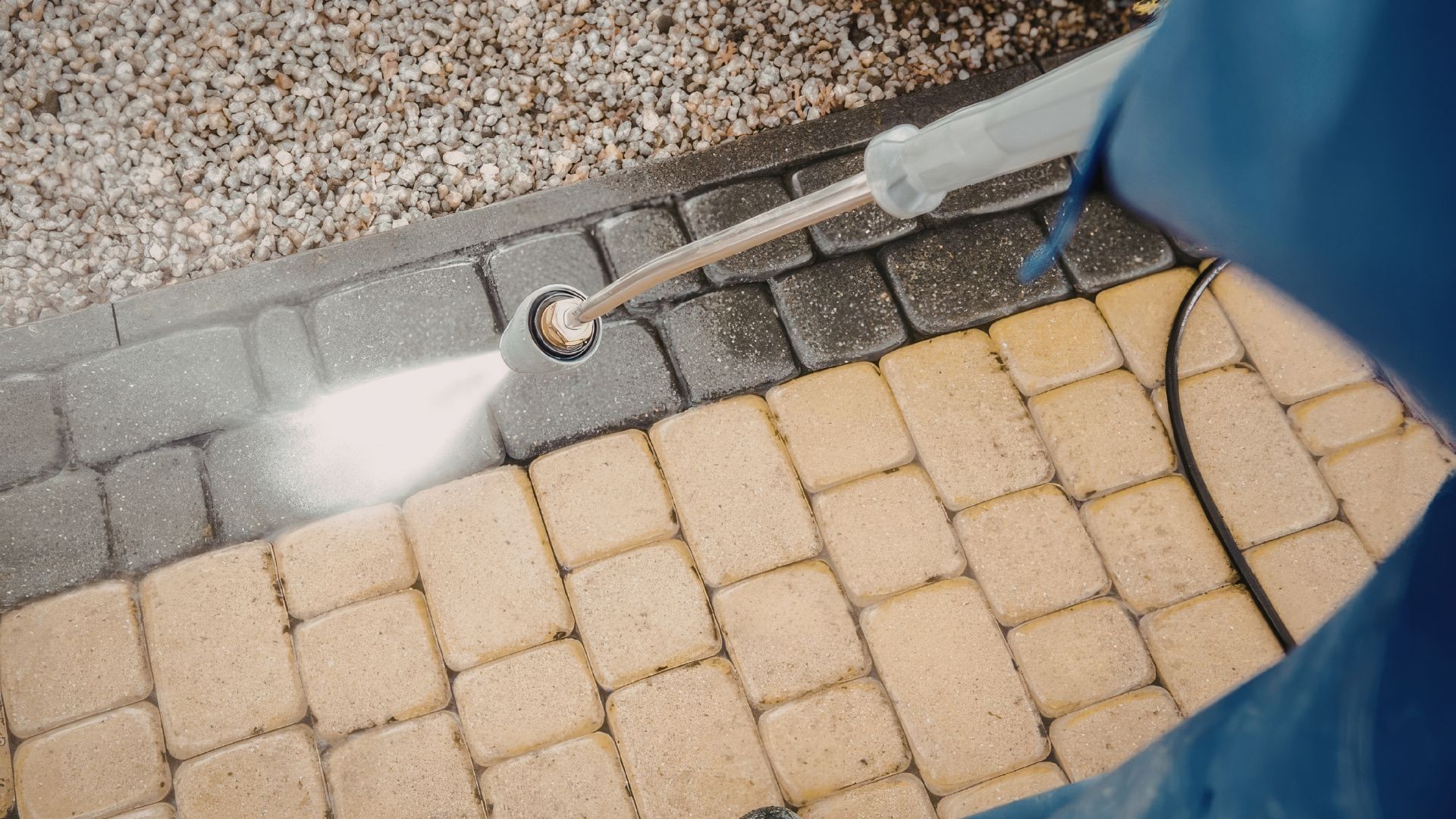 Pressure washing concrete pavers beside a gravel edge, with a spray wand and blue tarp visible.