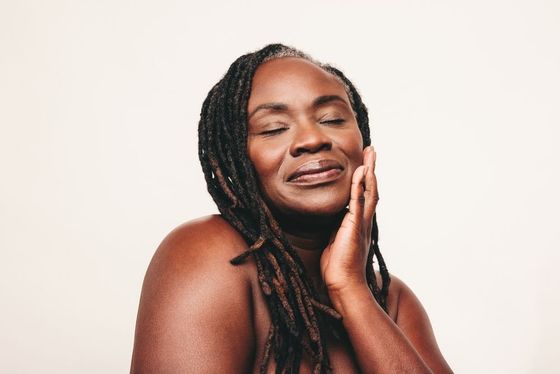 A woman with dreadlocks is smiling with her eyes closed and touching her face