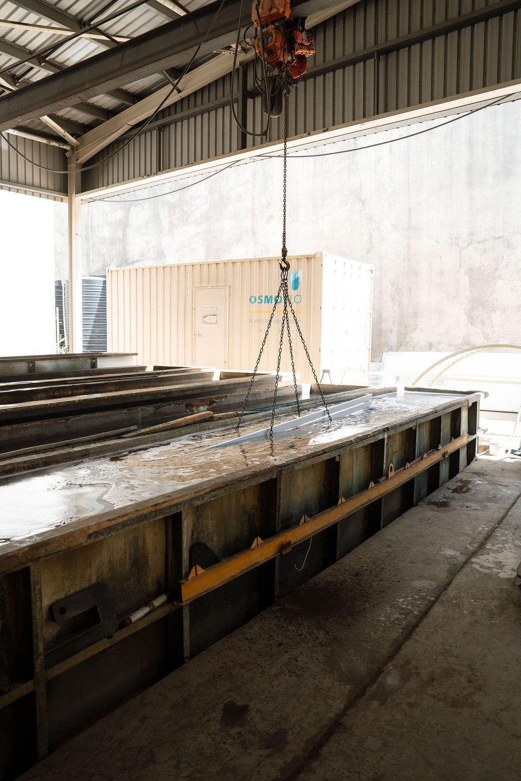 A crane chain suspends a metal object over a long industrial trough containing liquid inside a warehouse facility.