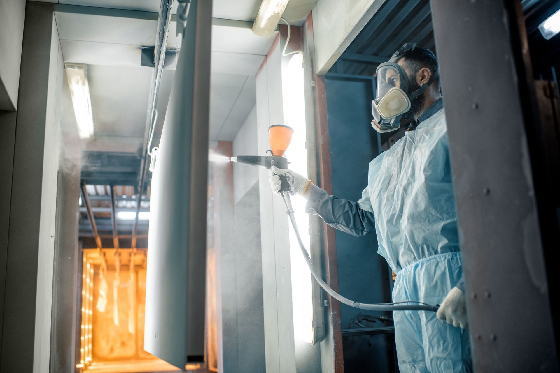 A worker wearing protective gear is performing a powder coating service using a spray gun.