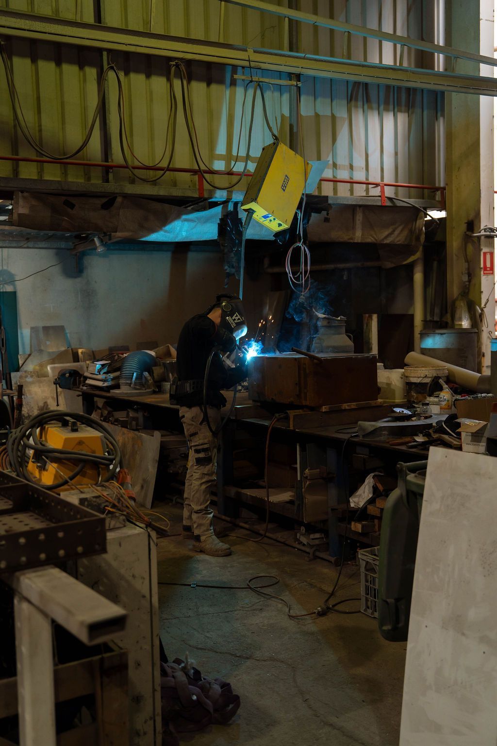 Welder working in a metal shop, sparks flying from the weld. A yellow overhead crane is visible.