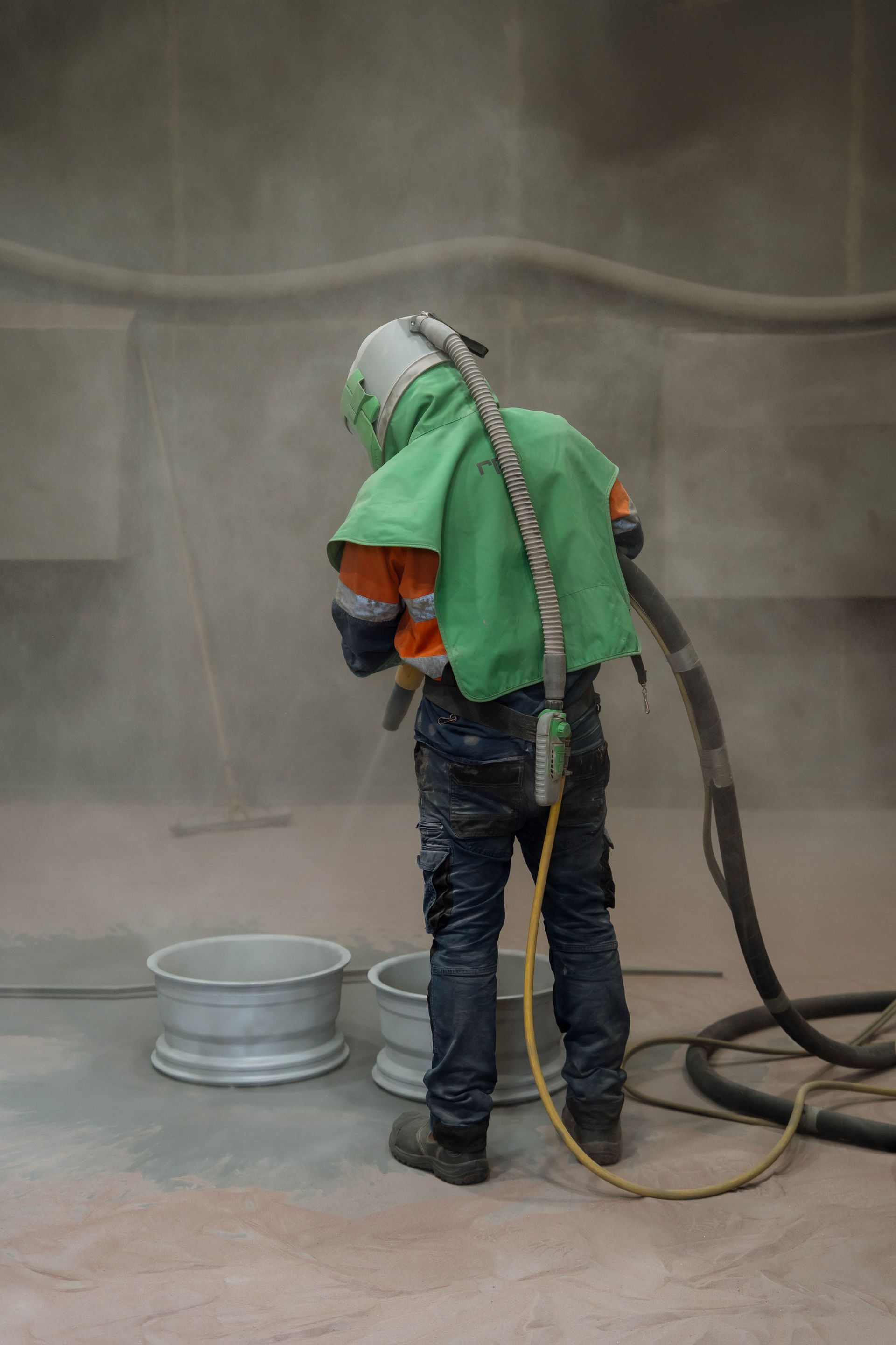 Person in protective suit sandblasting two car rims in a dusty workshop.