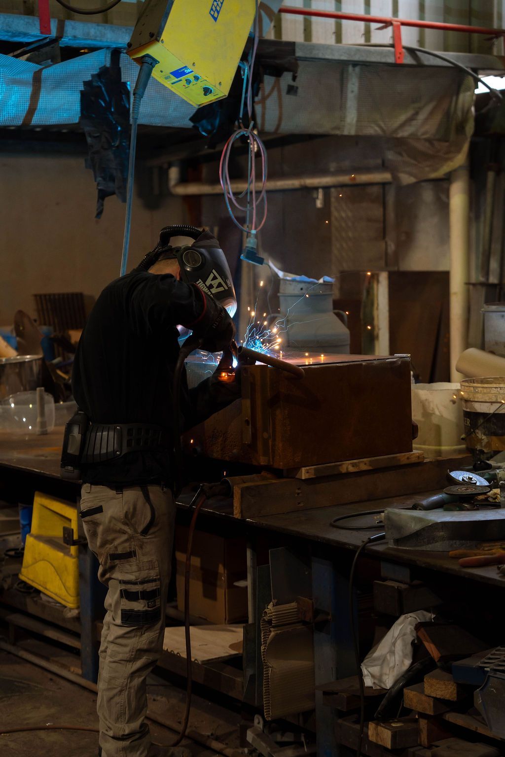 A person welds metal in a workshop, sparks flying. They wear a helmet and protective gear.