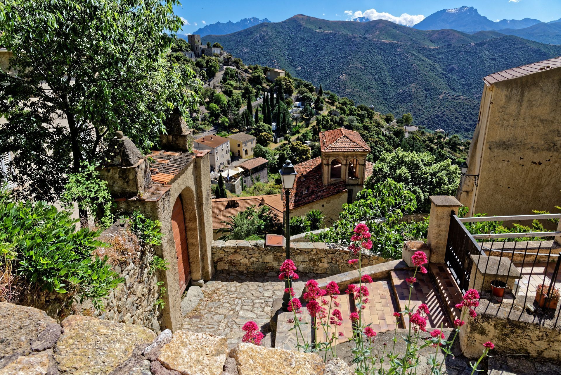 Eine Treppe führt hinauf zu einem kleinen Dorf mit Bergen im Hintergrund.