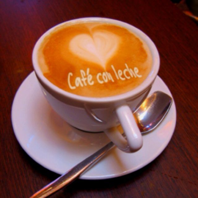 Café con leche in white cup on saucer, heart-shaped foam, spoon. “Cafe con leche” written on top.