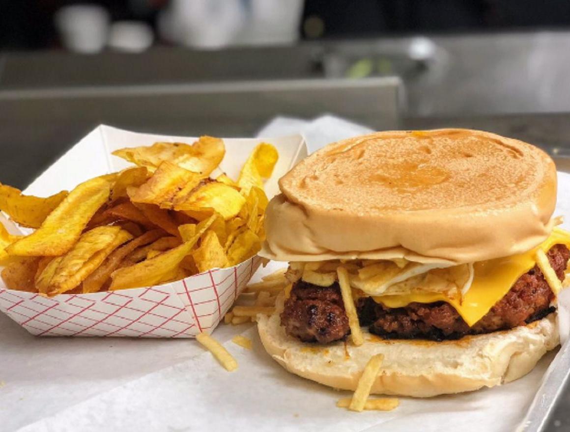 Burger with cheese and fried egg, served with plantain fries in a paper container.