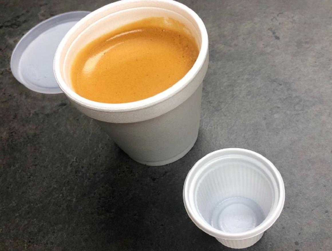 Coffee in a white foam cup, lid off, with an empty smaller cup on a gray surface.
