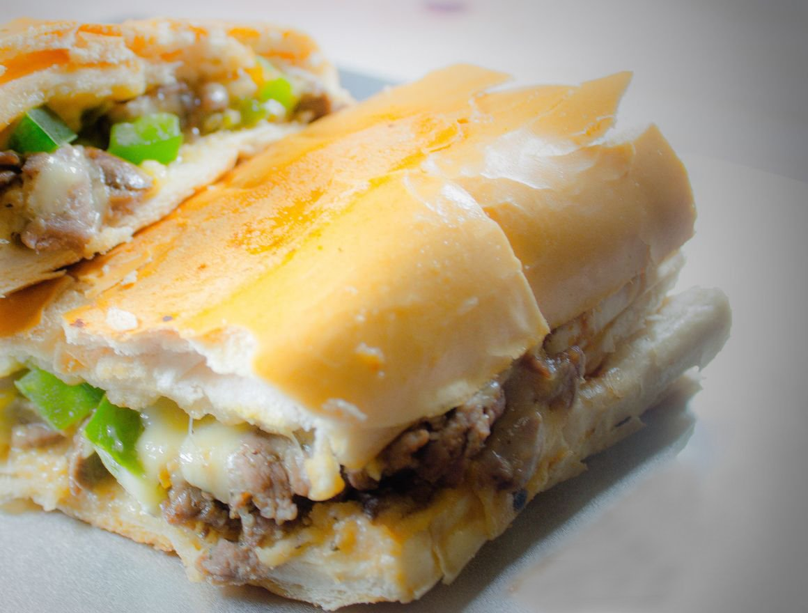 Philly cheesesteak sandwich on a white plate, cut in half, with visible melted cheese and green peppers.
