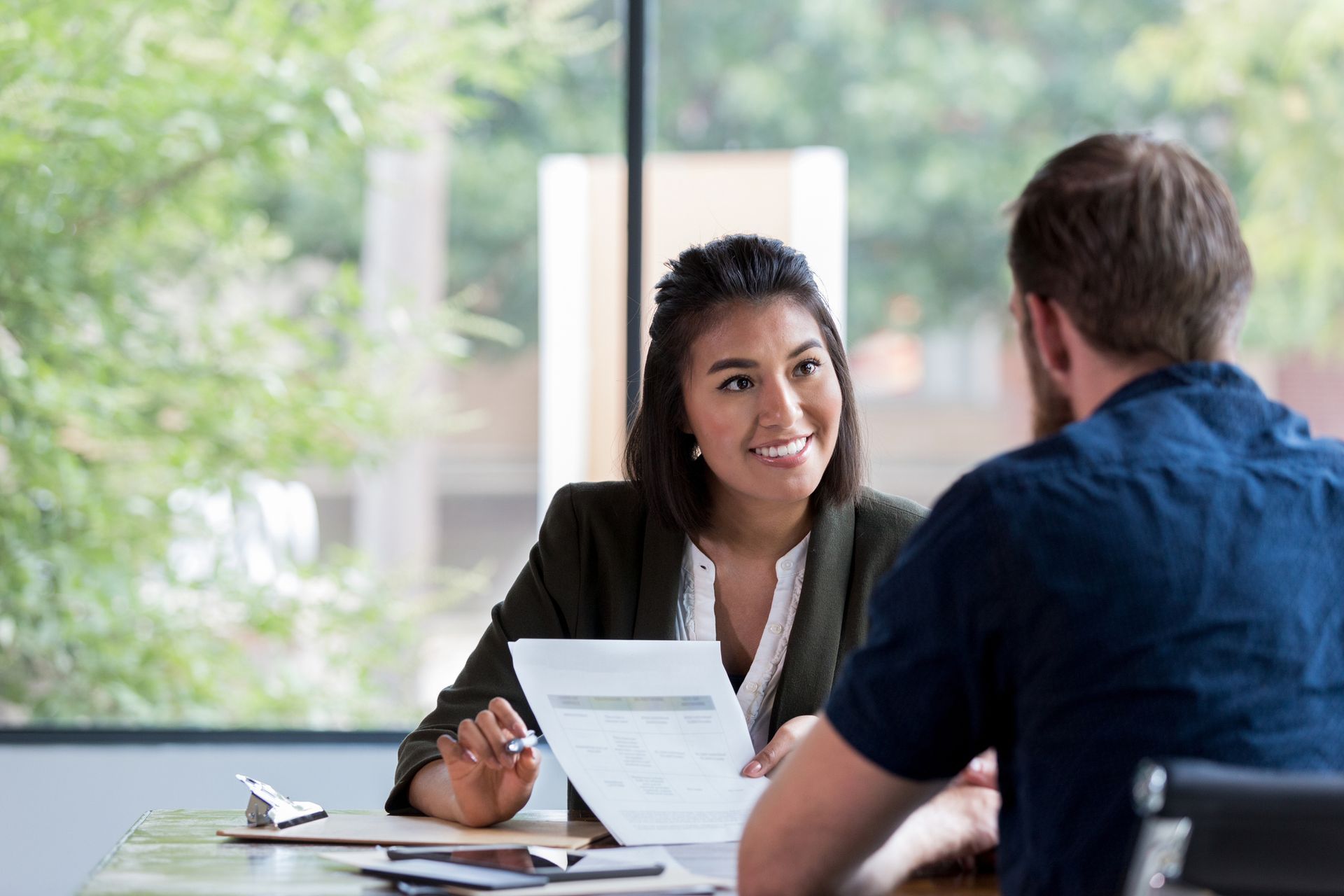 Woman in blazer smiles, reviewing document with a man at a table near a window.