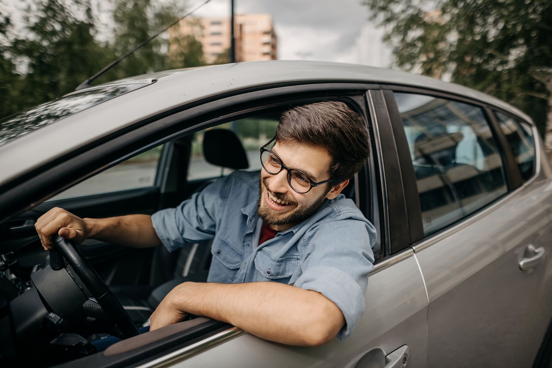 Man in glasses smiling, leaning out of car window, holding steering wheel.