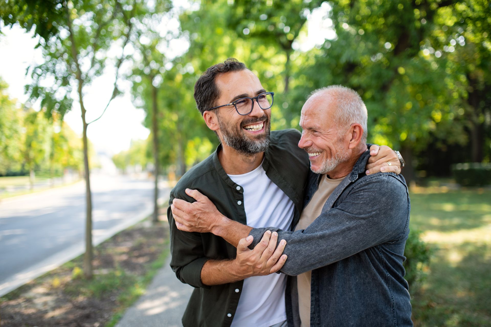 Two men, embracing and smiling on a tree-lined street.