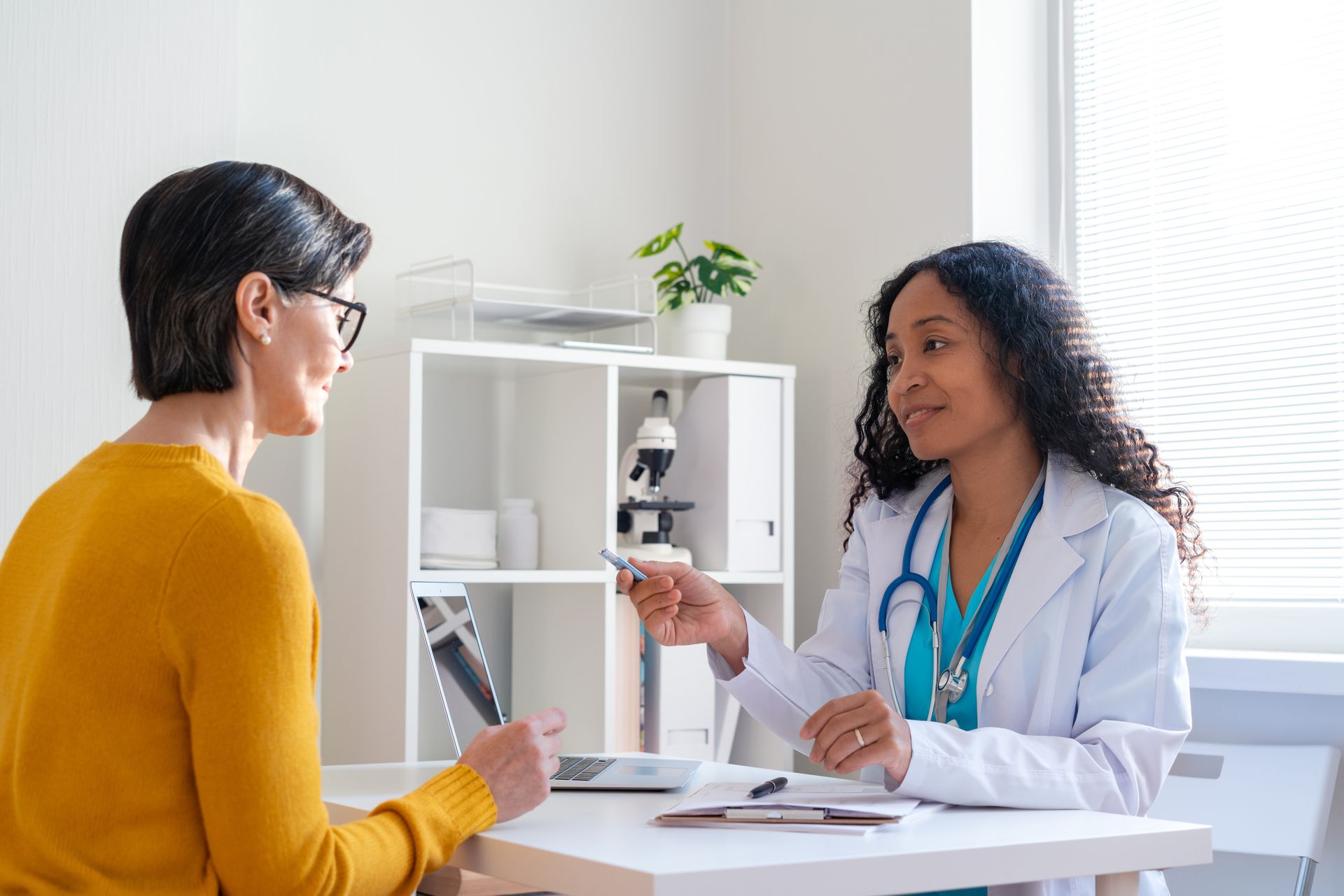 Doctor in white coat consults with patient in yellow sweater at a desk, light-filled office.