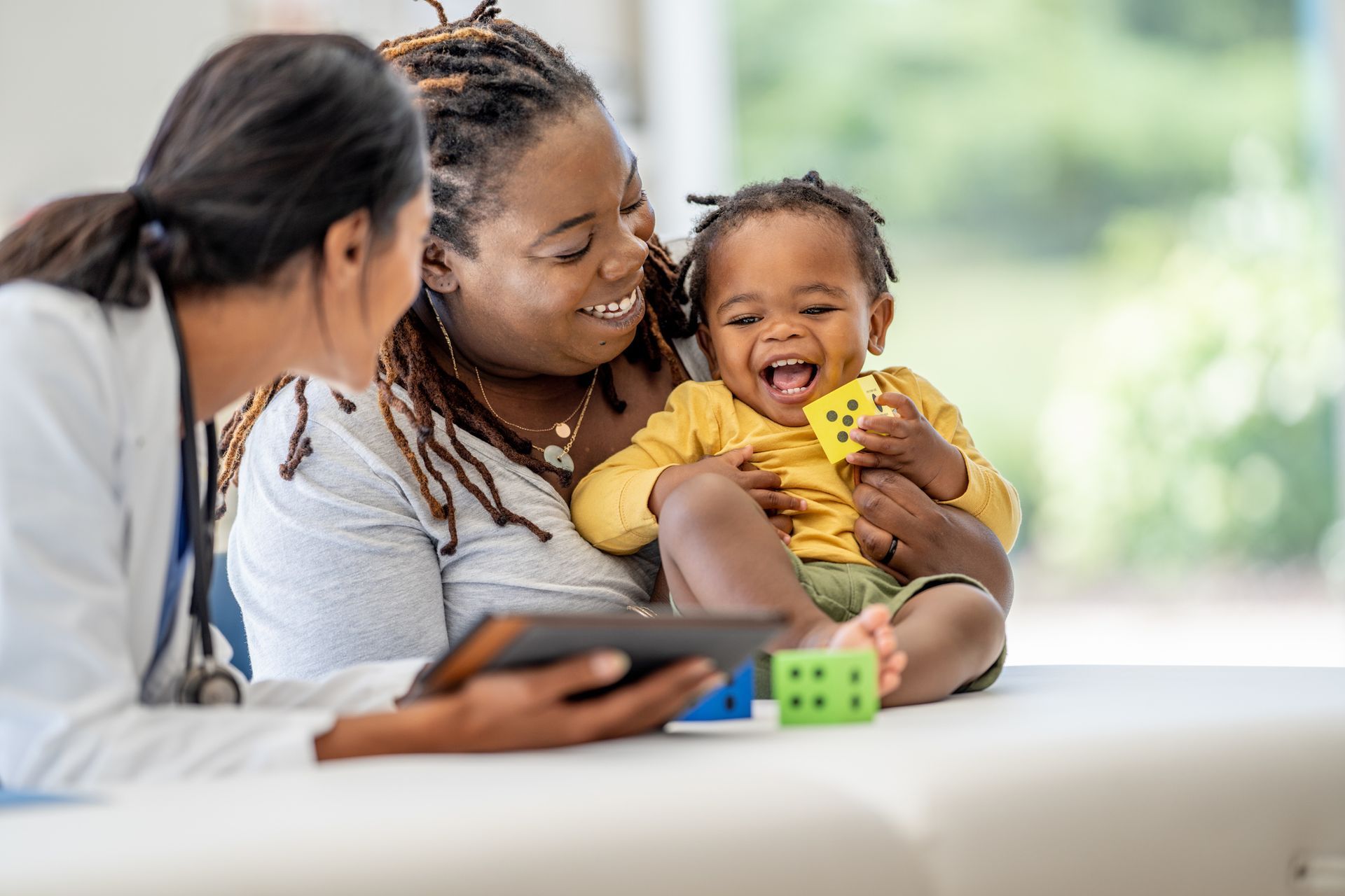 Doctor, parent, and baby; child laughs, holding toy, doctor looking at tablet, bright setting.